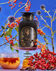 A black jar labeled "Magic Hour Virgo Tea of Virtue, Wit & Meticulous Magic" sits on a glass stand among vibrant orange berries, purple thistle flowers, and leafy branches under a blue sky. Beside it, a clear glass mug of Organic Tea with loose leaf tea and a cluster of red berries complete the harmonious arrangement.