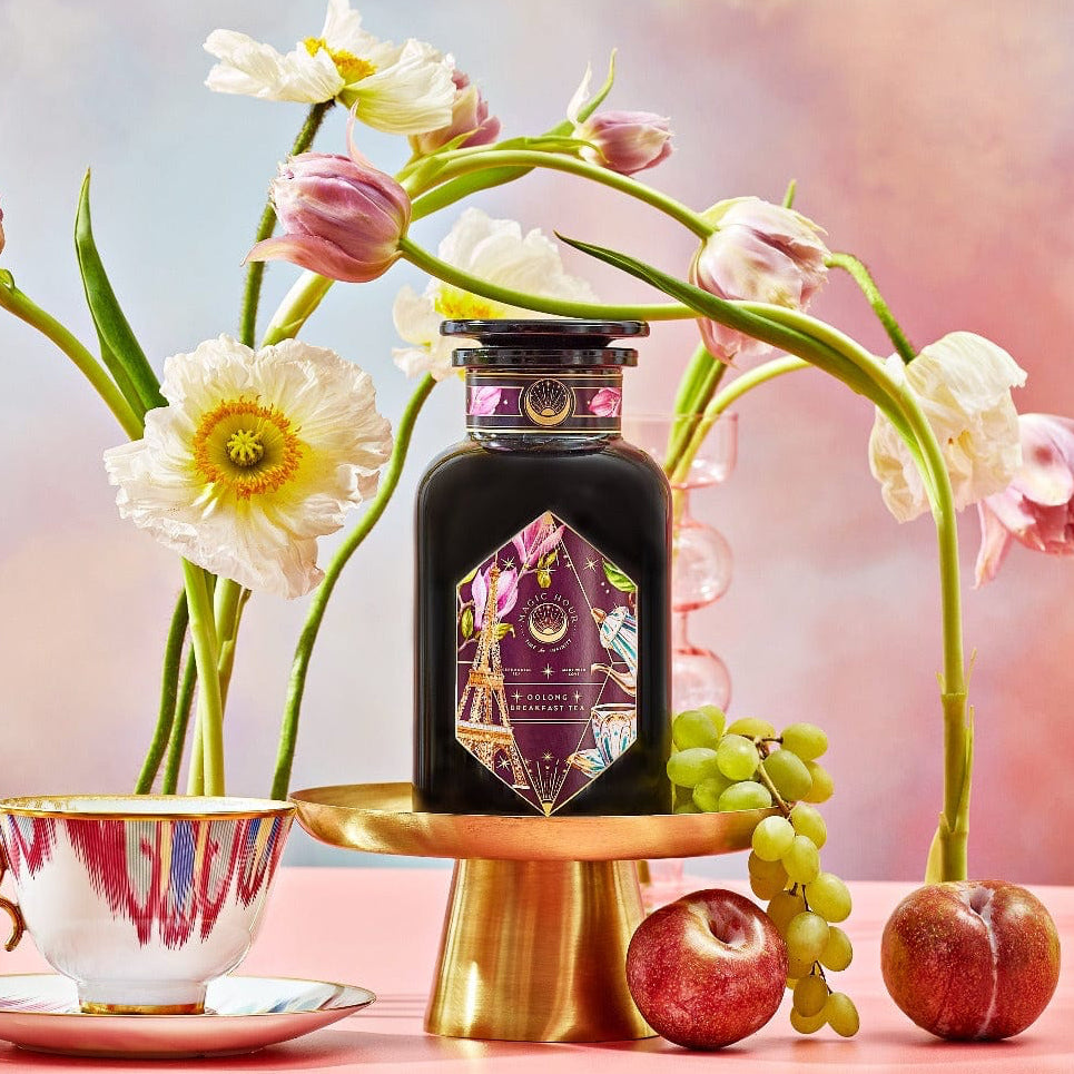 A bottle of "Breakfast in Paris-Oolong Black Tea Scented with Fruits & Flowers of Secret Parisian Gardens" is elegantly showcased on a gold stand, accompanied by flowers, fruits, and a teacup with a saucer. The background captures the essence of secret Parisian gardens, featuring a pastel gradient of soft pink, blue, and yellow tones.