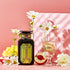 A bottle of Magic Hour's Peridot: Green Guayusa Oolong Mango-Peach Tea stands next to a wine glass with a flower and a golden strainer, all set against a pink background. White flowers, organic peach slices, and chunks of organic mango enhance the elegant display.