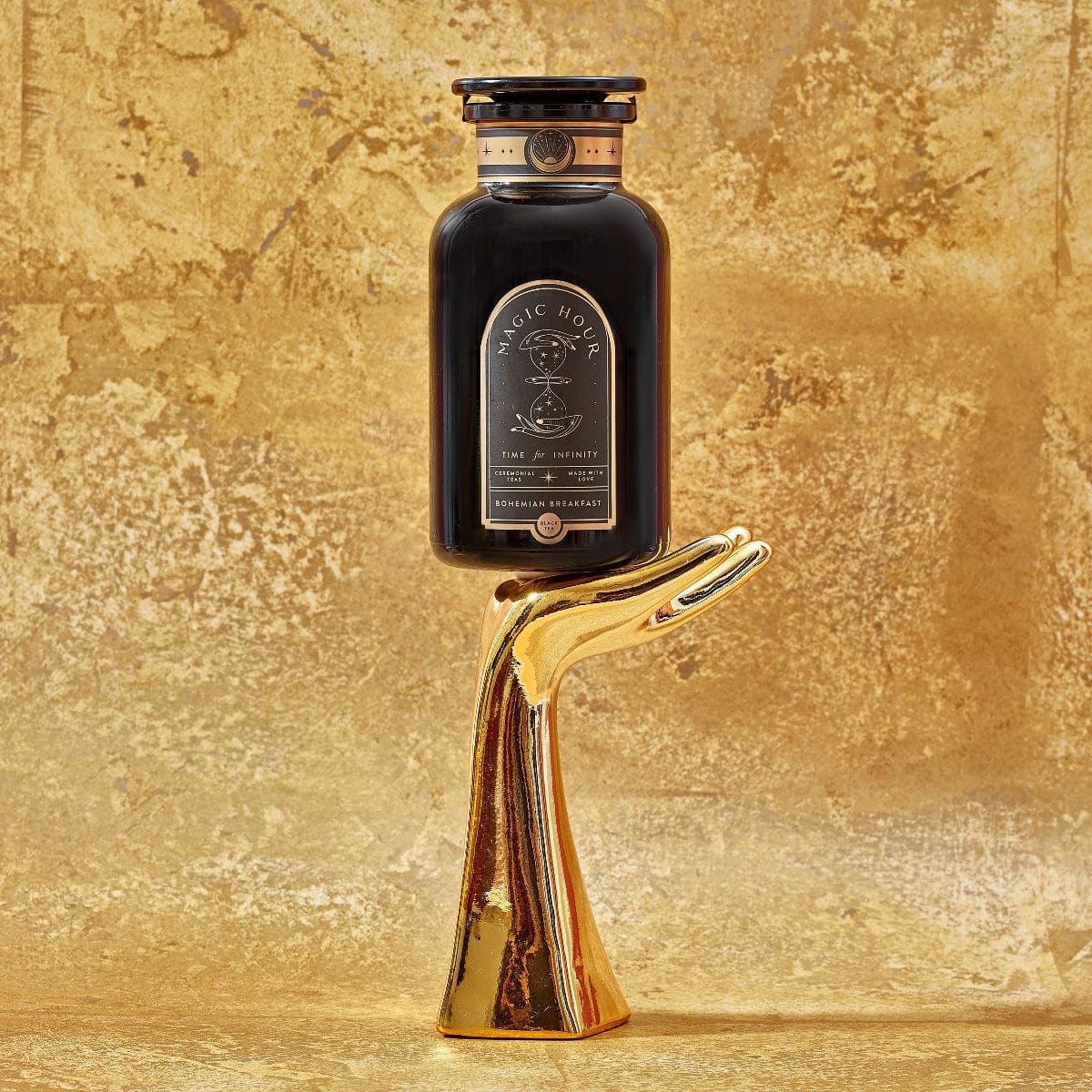 A dark-colored bottle labeled "[TEST] Bohemian Breakfast"—a distinctive blend of probiotic-rich black tea designed for energy, clarity, and digestion—rests on a gold, hand-shaped stand against a textured gold background. The bottle displays intricate details, instilling an air of luxury and elegance in this exquisite tableau.