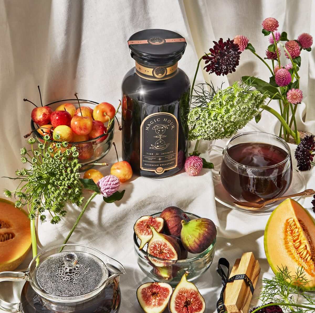 A bottle of herbal tonic is elegantly displayed among an assortment of figs, cherries, melon slices, and flowers on a white surface. To complete the vibrant, rustic setting, a glass teapot features [TEST] Bohemian Breakfast—Probiotic-Rich Black Tea for Energy, Clarity, Digestion—and next to it sits a cup filled with its dark and enriching brew.