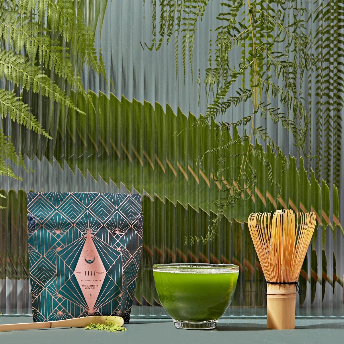 A serene setup featuring a bamboo whisk, a green bowl of matcha tea, and a beautifully designed package of Magic Hour Matcha 1111: Highest Grade of Ceremonial Matcha. The background features glass with a leafy, fern-like pattern, creating a calming, nature-inspired ambiance perfect for enjoying organic tea in your own magic hour.