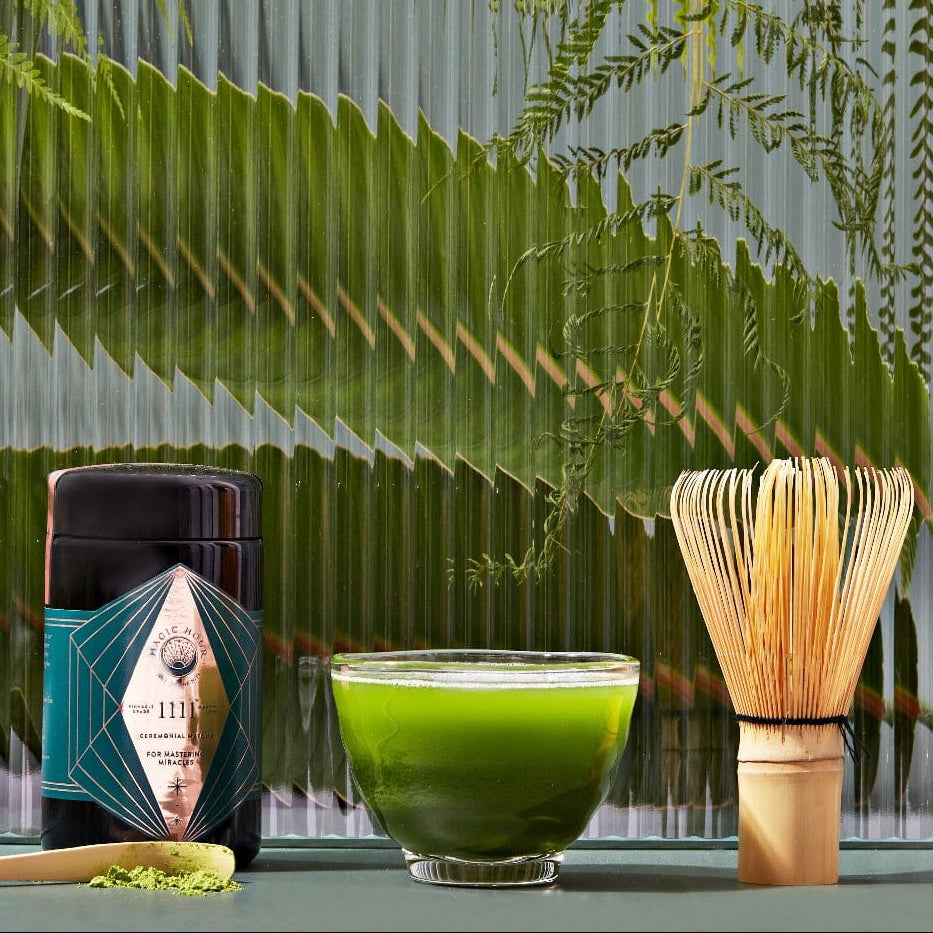 A Matcha 1111 : Highest Grade of Ceremonial Matcha kit on a green surface with a background of ferns. It includes a matcha container, a glass bowl with matcha tea, a bamboo whisk, and a small wooden spoon with matcha powder. The scene has a calm and natural ambiance, perfect for enjoying Magic Hour's organic tea selection.