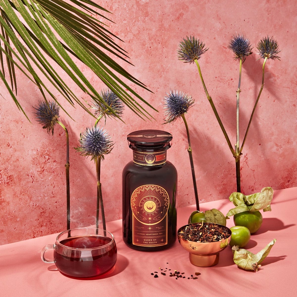 A dark bottle with a sun emblem stands on a pink surface surrounded by exotic flowers, green fruits, a teacup filled with a dark red liquid, and a bowl of organic loose leaf tea. A large green leaf dangles from above, set against a textured pink background. Magic Hour Queen of the South: Delicious Cocoa Detox Tea awaits your senses.
