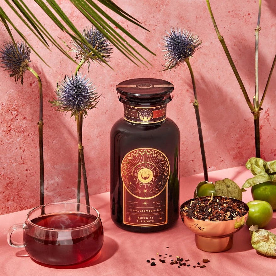 A dark bottle with a sun emblem stands on a pink surface surrounded by exotic flowers, green fruits, a teacup filled with a dark red liquid, and a bowl of organic loose leaf tea. A large green leaf dangles from above, set against a textured pink background. Magic Hour Queen of the South: Delicious Cocoa Detox Tea awaits your senses.