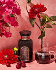 A vibrant still life scene features a bottle of Ruby Moon™ : Hibiscus Elderberry Tea by Club Magic Hour, a steaming transparent teacup, a bunch of fresh cherries, and bright red flowers against a pink backdrop. The arrangement highlights the rich red hues and the elegance of the tea setting.