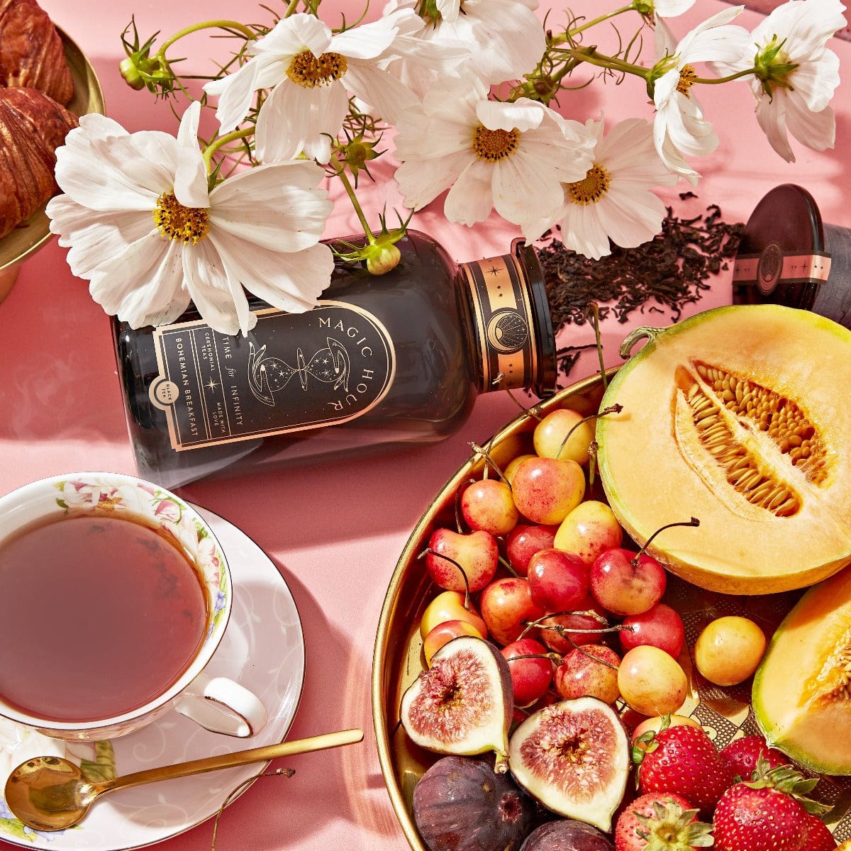 A vibrant arrangement featuring a tea set with a cup of [TEST] Bohemian Breakfast - Probiotic-Rich Black Tea for Energy, Clarity, Digestion, accompanied by a jar of magic hour tea, a plate of fruit including cherries, figs, melon, and strawberries, and a bouquet of white flowers, all beautifully displayed on a pink tablecloth.