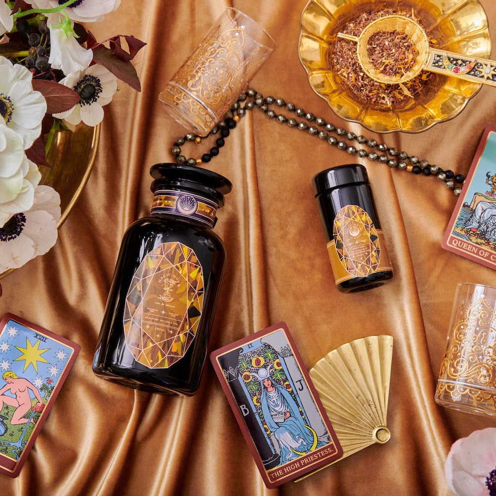 An elegant display featuring tarot cards, a black and gold bottle, glasses, a fan, and a floral arrangement on a silky, golden fabric. The tarot cards are arranged around the bottle, lending a mystical and luxurious ambiance to the setting—perfect for cozy sips of Magic Hour's Topaz - Caramel Apple Oolong Tea from the case packs.