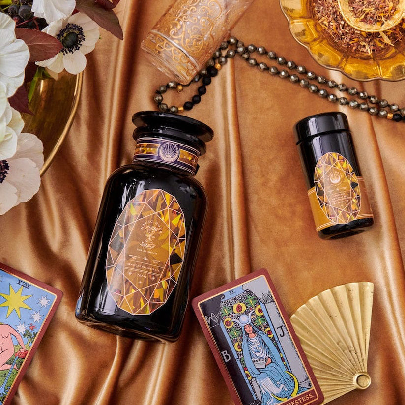 An elegant display featuring tarot cards, a black and gold bottle, glasses, a fan, and a floral arrangement on a silky, golden fabric. The tarot cards are arranged around the bottle, lending a mystical and luxurious ambiance to the setting—perfect for cozy sips of Magic Hour's Topaz - Caramel Apple Oolong Tea from the case packs.