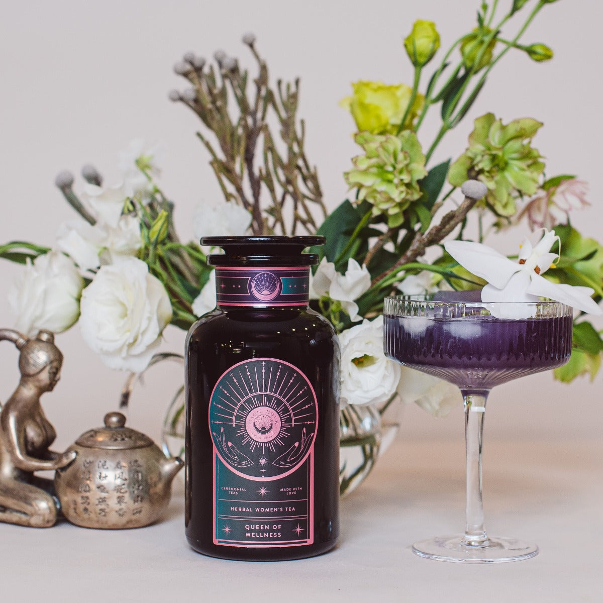 A glass bottle labeled 'Magic Hour Queen of Wellness: Women's Hormone Balancing Tea for PMS, Healthy Cycles & Menopause' with a dark brownish-purple liquid stands next to a glass goblet of the same liquid garnished with a white flower. In the background are white and green flowers, a small statue, and a teapot alongside an assortment of loose leaf tea from Magic Hour.