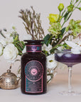 A glass bottle labeled "Magic Hour Queen of Wellness: Women's Hormone Balancing Tea for PMS, Healthy Cycles & Menopause" with a dark brownish-purple liquid stands next to a glass goblet of the same liquid garnished with a white flower. In the background are white and green flowers, a small statue, and a teapot alongside an assortment of loose leaf tea from Magic Hour.