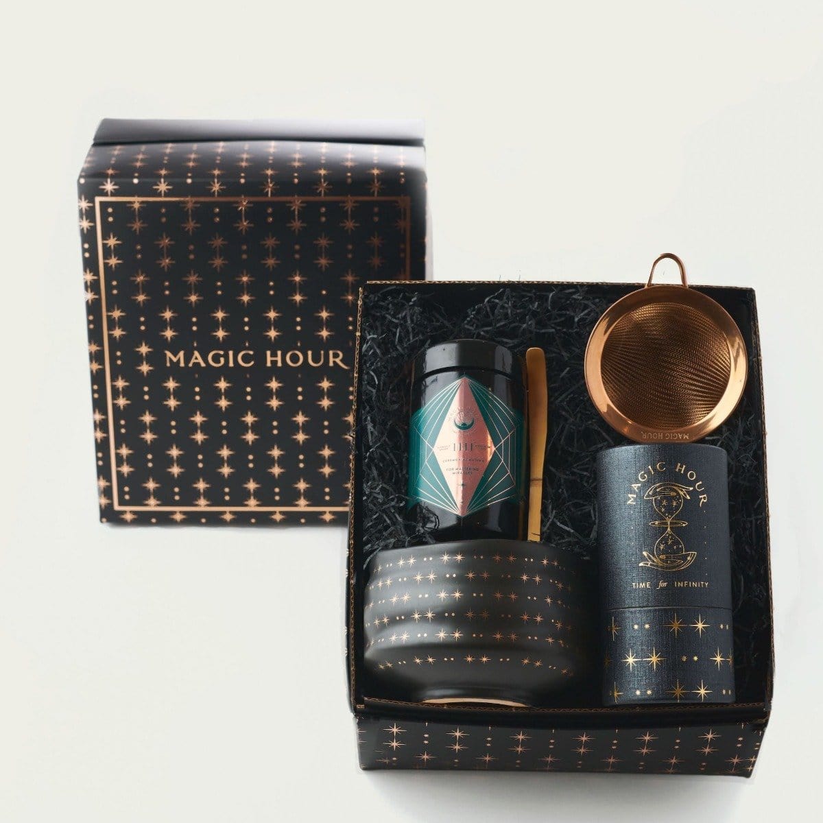 A beautifully packaged tea gift set named "Matcha 1111 : Gift Set" from Magic Hour contains a tin of loose leaf tea, a tea strainer, and a jar with a spoon, all nestled in a black box with gold star patterns. The box lid sits next to the set, flaunting the same design and "MAGIC HOUR TEA" text.