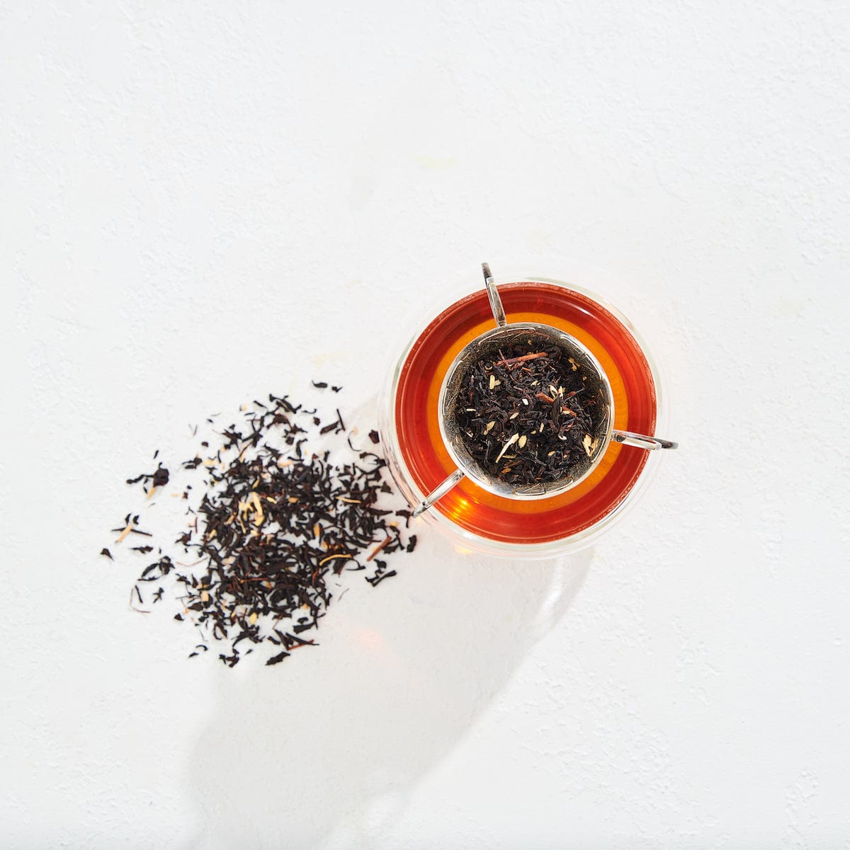 A cup filled with amber-colored Virgo Tea of Virtue, Wit &amp; Meticulous Magic sits on a white surface with a tea strainer containing loose tea leaves resting on top. To the left of the cup, a small pile of loose leaf tea is scattered on the surface. The scene is brightly lit, evoking a serene Magic Hour moment.