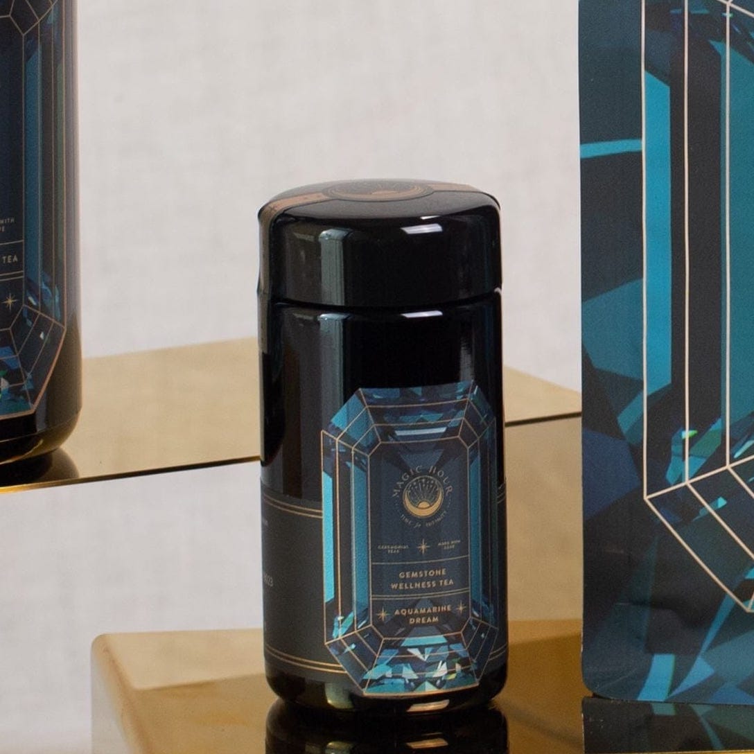 A black cylindrical container with a blue geometric design sits on a golden reflective shelf. The label reads &quot;Magic Hour&quot; with &quot;Aquamarine Dream - Soothing Herbal Ayurvedic Adrenal Tonic&quot; written below. This loose leaf tea is flanked by a similar design on the right, adding to the allure of Magic Hour Tea.