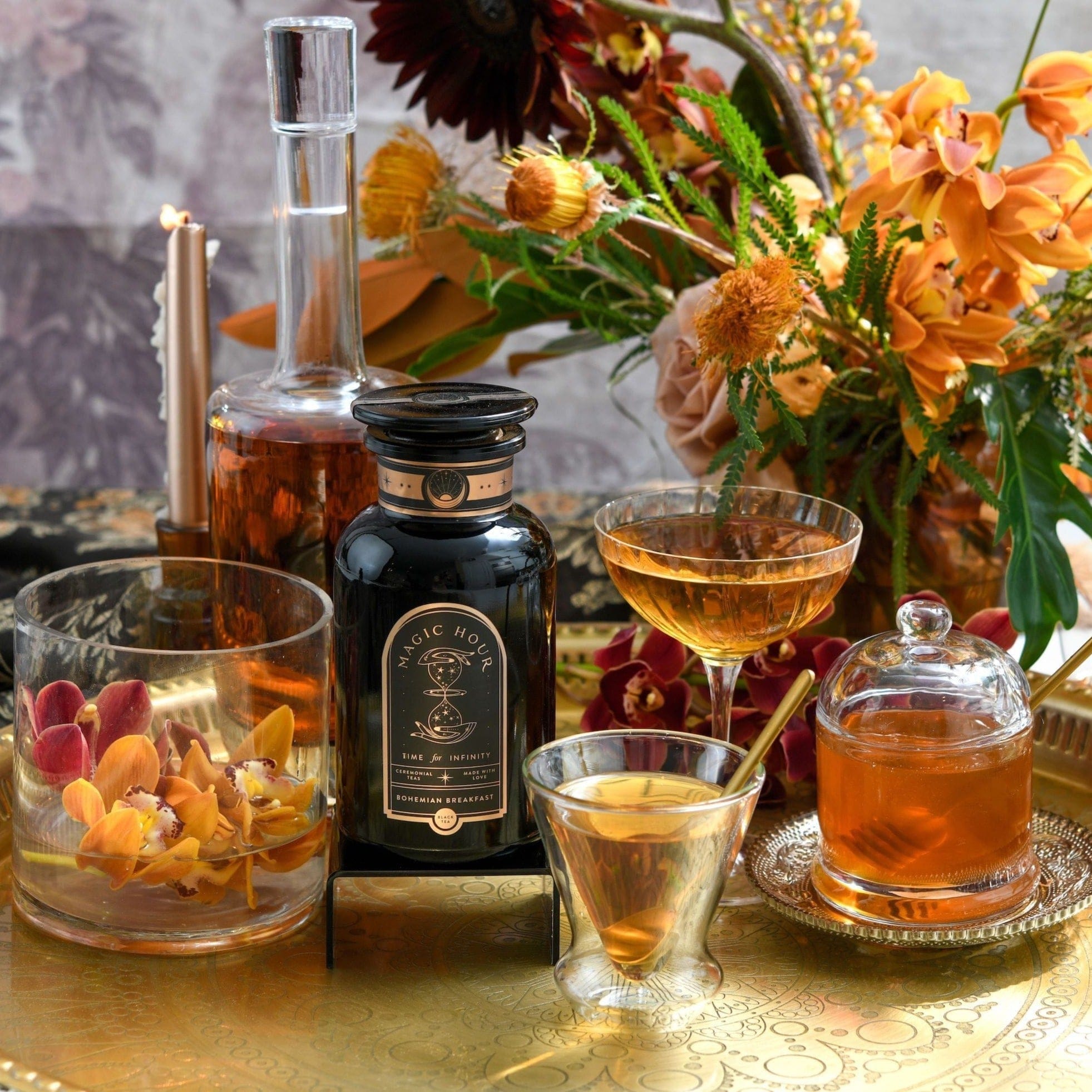 A decorative setup featuring bottles and glasses with amber liquids, surrounded by vibrant orange and yellow flowers on a gold tray, showcases the elegance of the [TEST] Bohemian Breakfast – a probiotic-rich black tea designed for energy, clarity, and digestion. This arrangement creates a warm and inviting atmosphere.