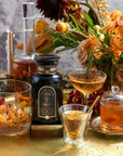 A decorative setup featuring bottles and glasses with amber liquids, surrounded by vibrant orange and yellow flowers on a gold tray, showcases the elegance of the [TEST] Bohemian Breakfast – a probiotic-rich black tea designed for energy, clarity, and digestion. This arrangement creates a warm and inviting atmosphere.