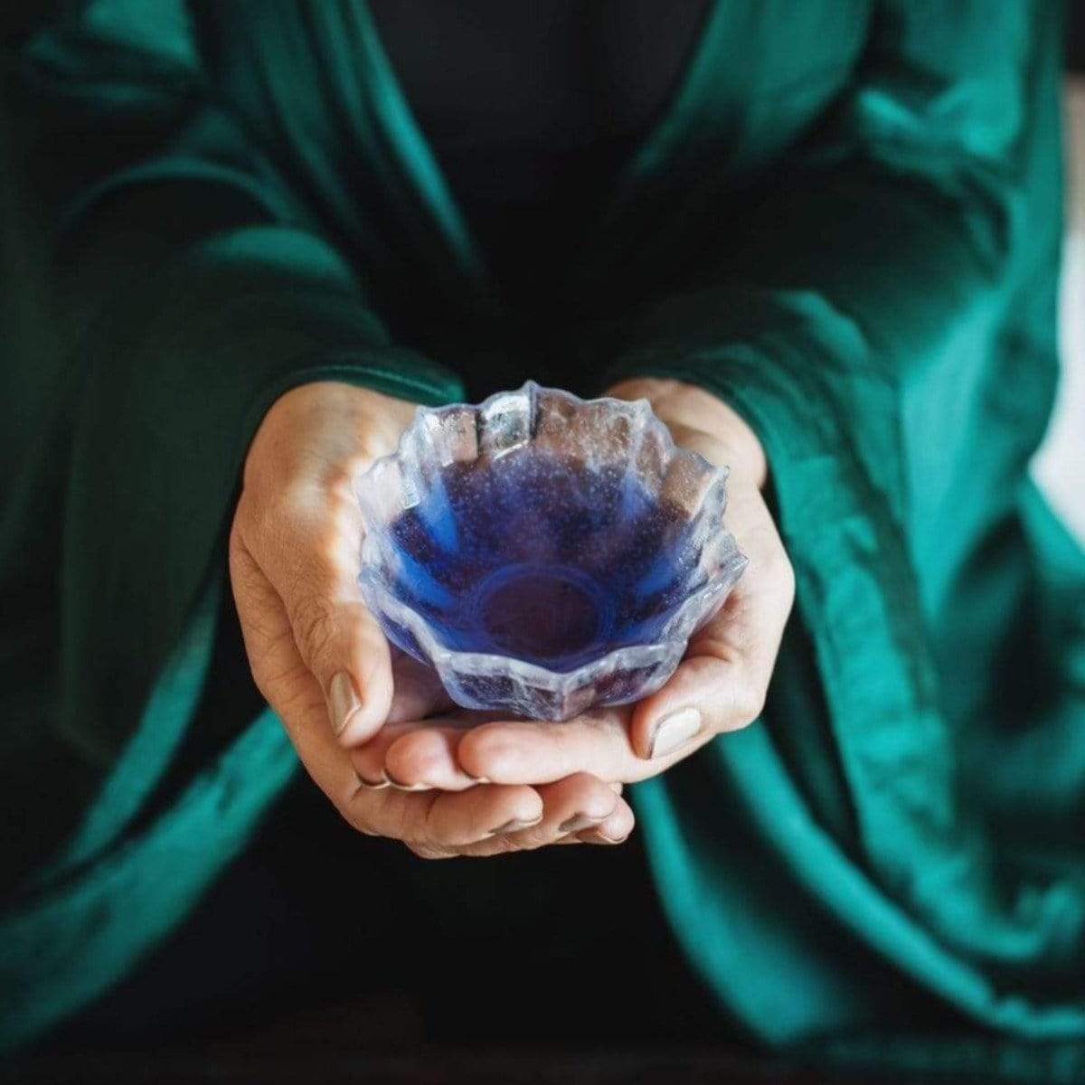 A pair of hands cradles a small, blue, leaf-shaped bowl with a person in the background dressed in a dark green robe. The bowl's contents hint at the vibrant tones of the Butterfly Pea Flower: Finely Cut- ARCHIVE product.
