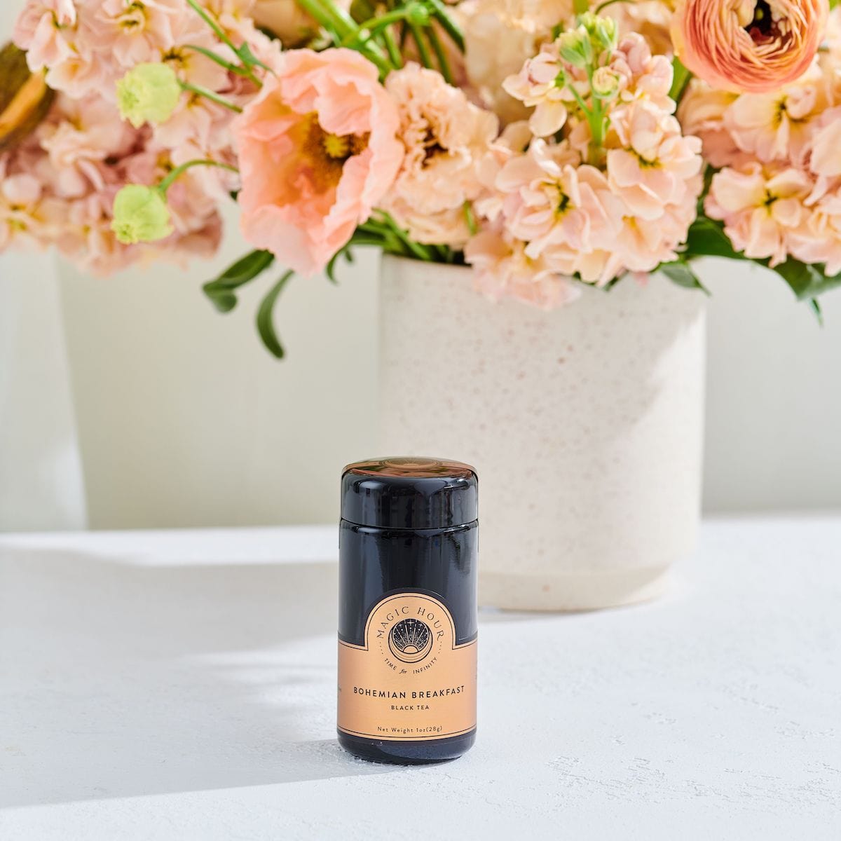 A jar of [TEST] Bohemian Breakfast- Probiotic-Rich Black Tea for Energy, Clarity, Digestion is placed on a white table in front of a bouquet of pink and peach flowers in a white vase. The scene is bright and inviting.
