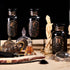 An arrangement of decorative items including three black glass jars labeled "Aries," "Taurus," and "Gemini." The scene features a brown teapot filled with Astrology Bundle: Aries, Taurus, Gemini - Magic Hour by Magic Hour, two glass perfume bottles, a chess piece-like statue, and various trinkets on a beige surface with a black background.