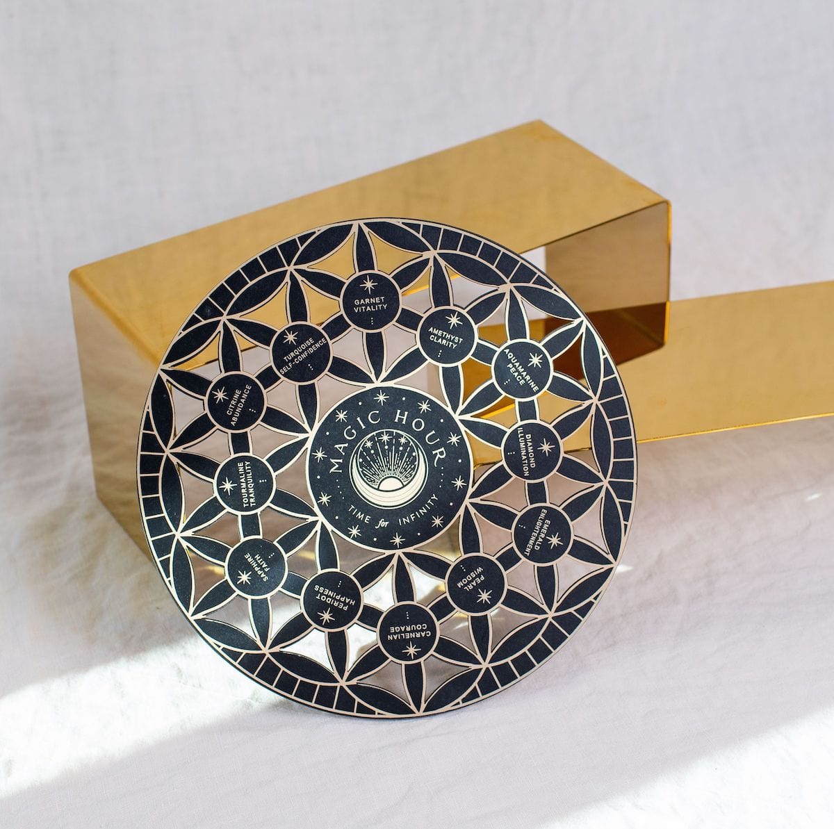 A round black and silver decorative plate with intricate geometric patterns and text &quot;Crystal Grid: Flower of Life Tea Ceremony Altar&quot; at the center, surrounded by smaller circles with text like &quot;gratitude,&quot; &quot;immunity,&quot; and &quot;healing.&quot; The plate is set against a white cloth backdrop, next to a golden rectangular object.