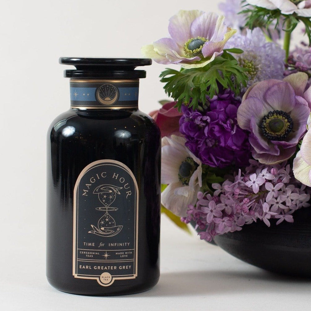 A black bottle labeled "Magic Hour - Earl Greater Grey: Tea for the Bright &amp; Bold" sits beside a vibrant bouquet of purple and white flowers. Featuring an apothecary design with a cosmic hourglass label, this mysterious vessel contains Magic Hour's finest loose leaf tea, and the flowers are arranged in a black vase.