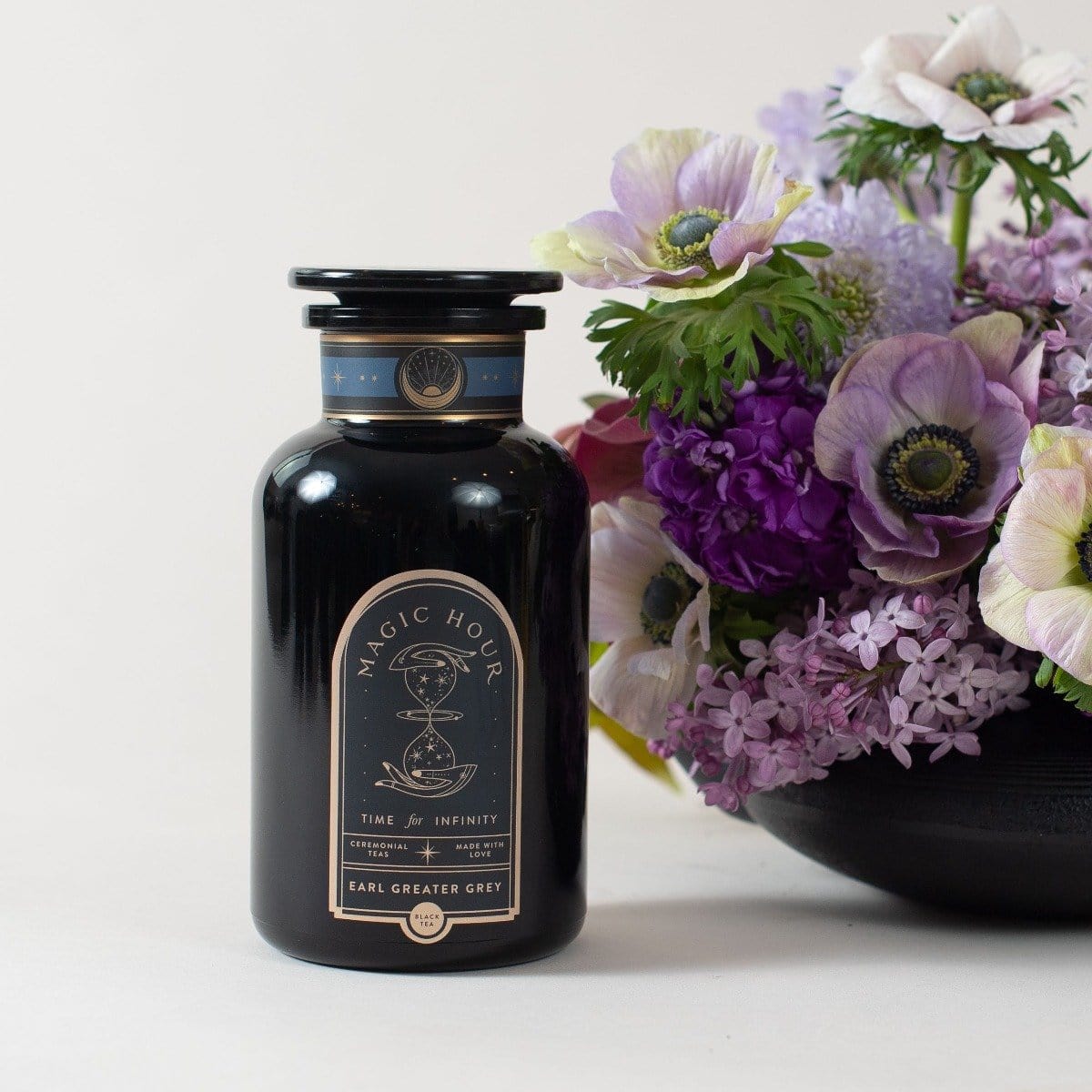A black bottle labeled "Magic Hour - Earl Greater Grey: Tea for the Bright & Bold" sits beside a vibrant bouquet of purple and white flowers. Featuring an apothecary design with a cosmic hourglass label, this mysterious vessel contains Magic Hour's finest loose leaf tea, and the flowers are arranged in a black vase.