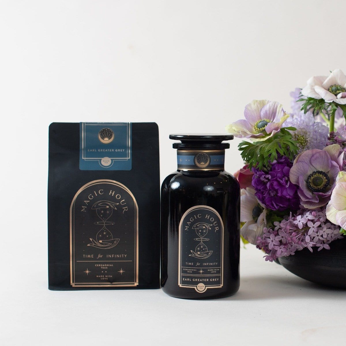 A black bag and a black jar of Magic Hour's "Earl Greater Grey: Tea for the Bright & Bold" loose leaf tea are placed next to a vase containing a bouquet of flowers with purple, white, and pink blooms. The packaging, which highlights the organic tea blend, features gold and white text with a mystical design.