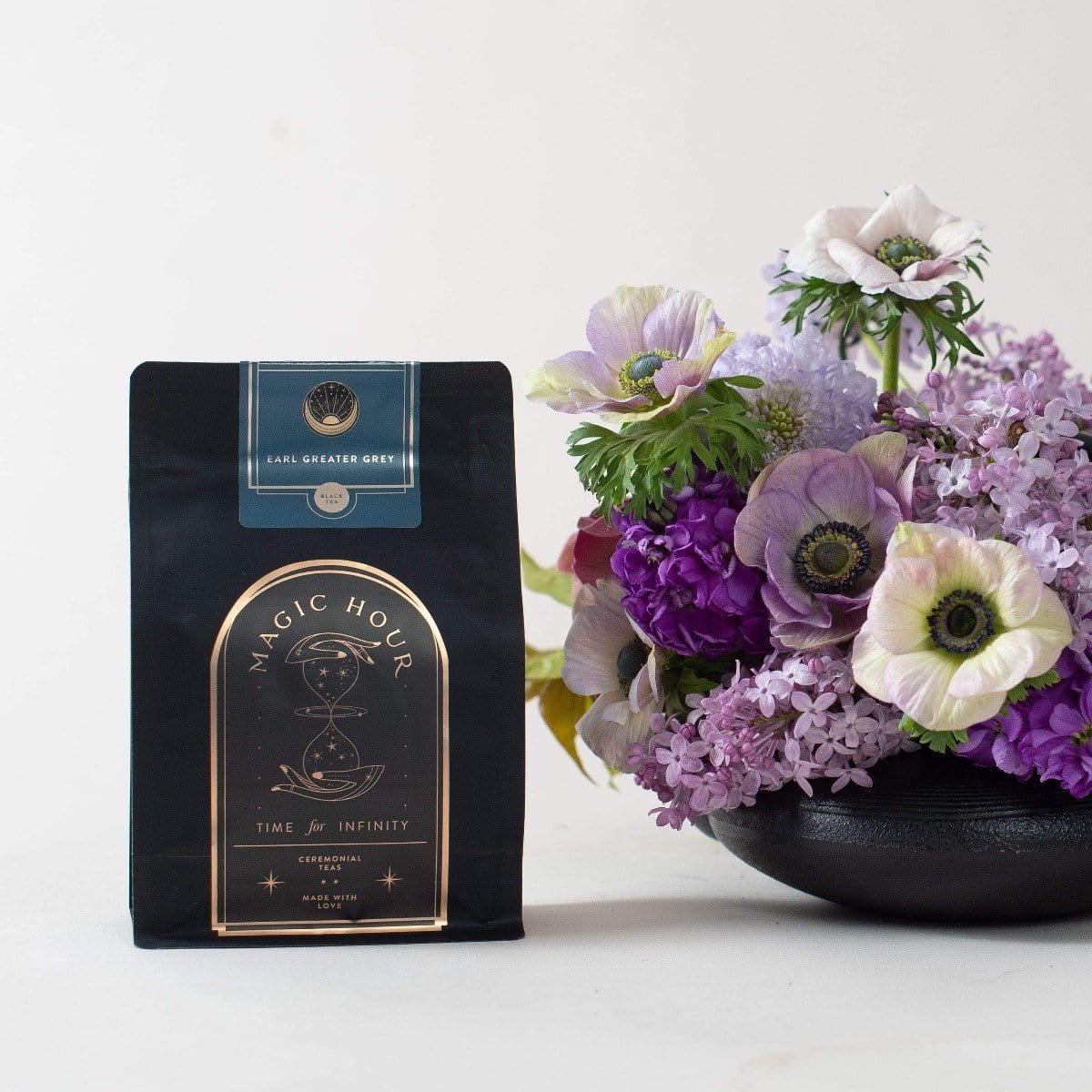 A black package of Magic Hour organic tea sits on a white surface next to a bouquet of purple and white flowers in a black vase. The tea package has a label that reads "Earl Greater Grey: Tea for the Bright & Bold" and "Time & Infinity" with an hourglass graphic, making this loose leaf tea experience timeless.