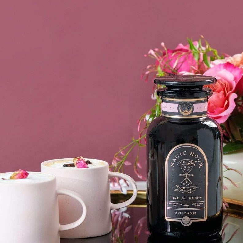 A bottle of Gypsy Rose Black Tea by Magic Hour sits on a round golden tray with a black base. Next to it are two white cups filled with tea, garnished with small rose petals. In the background is a bouquet of pink roses against a mauve wall.