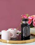 A bottle of Gypsy Rose Black Tea by Magic Hour sits on a round golden tray with a black base. Next to it are two white cups filled with tea, garnished with small rose petals. In the background is a bouquet of pink roses against a mauve wall.