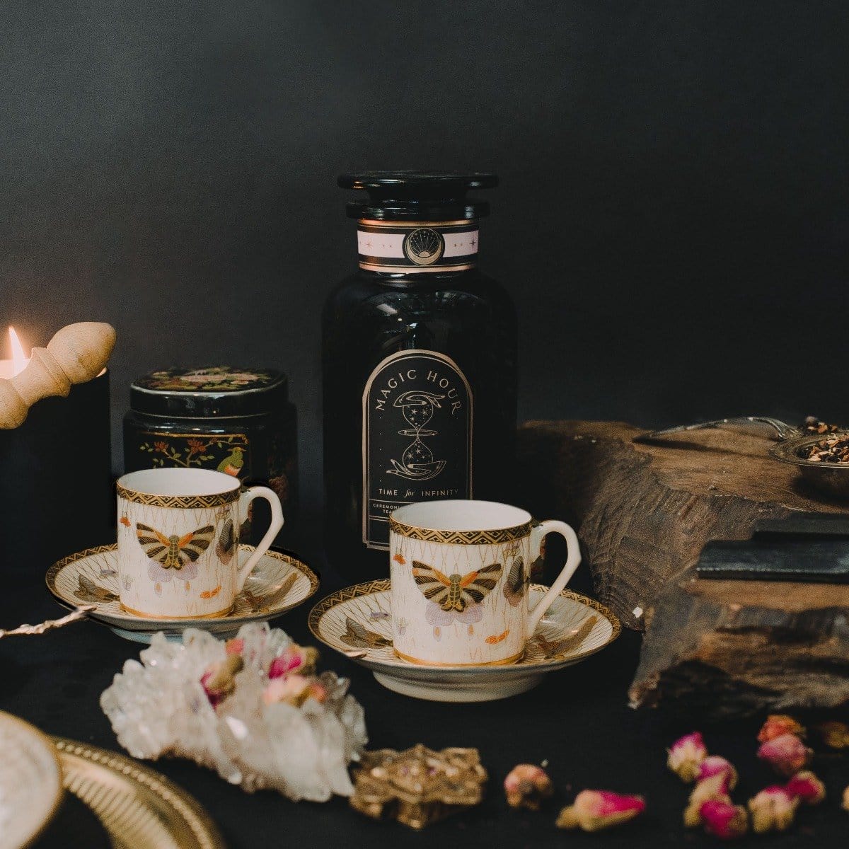 A beautifully styled scene features two ornate teacups and saucers adorned with a butterfly design. Behind the teacups is a dark bottle labeled "Gypsy Rose Black Tea" by Magic Hour and a lit candle. Other items include crystal and dried flowers, all set against a dark backdrop.