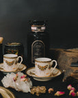 A beautifully styled scene features two ornate teacups and saucers adorned with a butterfly design. Behind the teacups is a dark bottle labeled "Gypsy Rose Black Tea" by Magic Hour and a lit candle. Other items include crystal and dried flowers, all set against a dark backdrop.