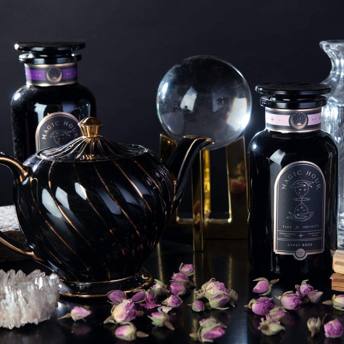 A dark, elegant display features two black jars labeled "Black Tea Magic Sampler Box- Bestselling Organic Black Teas for Energy & Vitality," a black teapot with gold accents, dried rosebuds, a crystal ball on a stand, a crystal cluster, and glassware against a dark backdrop. The scene exudes mystical and luxurious vibes with the allure of Magic Hour.
