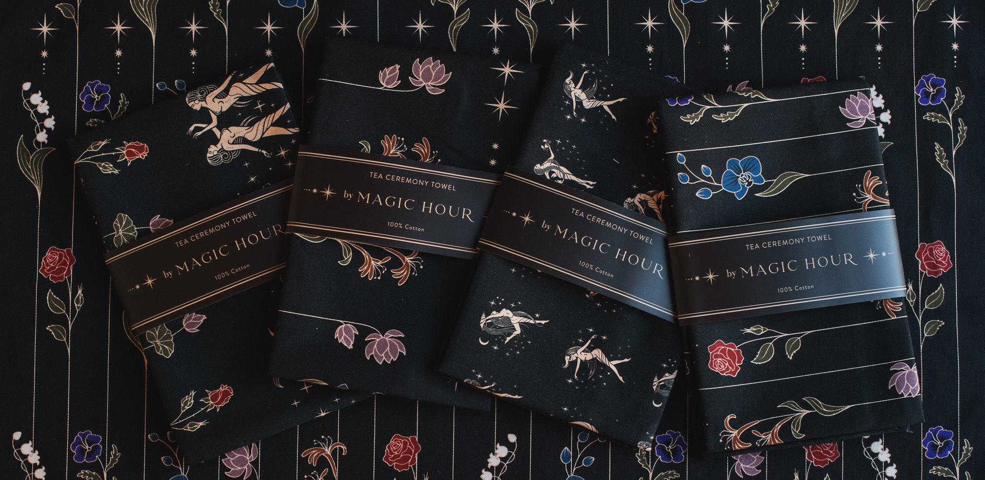 A display of four neatly folded Tea Ceremony Towels Bundle, each wrapped with a label reading "Magic Hour." The towels feature various floral and celestial designs in vibrant colors, placed on a fabric backdrop adorned with intricate floral and star patterns.