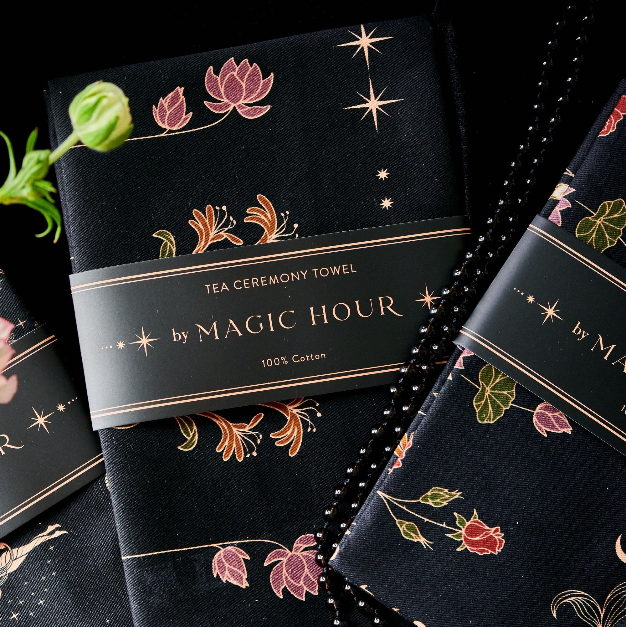 A close-up of the Tea Ceremony Towels Bundle by Magic Hour with floral and star patterns in gold and colorful designs. One towel is partially unwrapped, revealing its delicate design. Perfect for any Magic Hour Tea ritual, the packaging reads "TEA CEREMONY TOWELS BUNDLE by MAGIC HOUR 100% Cotton.