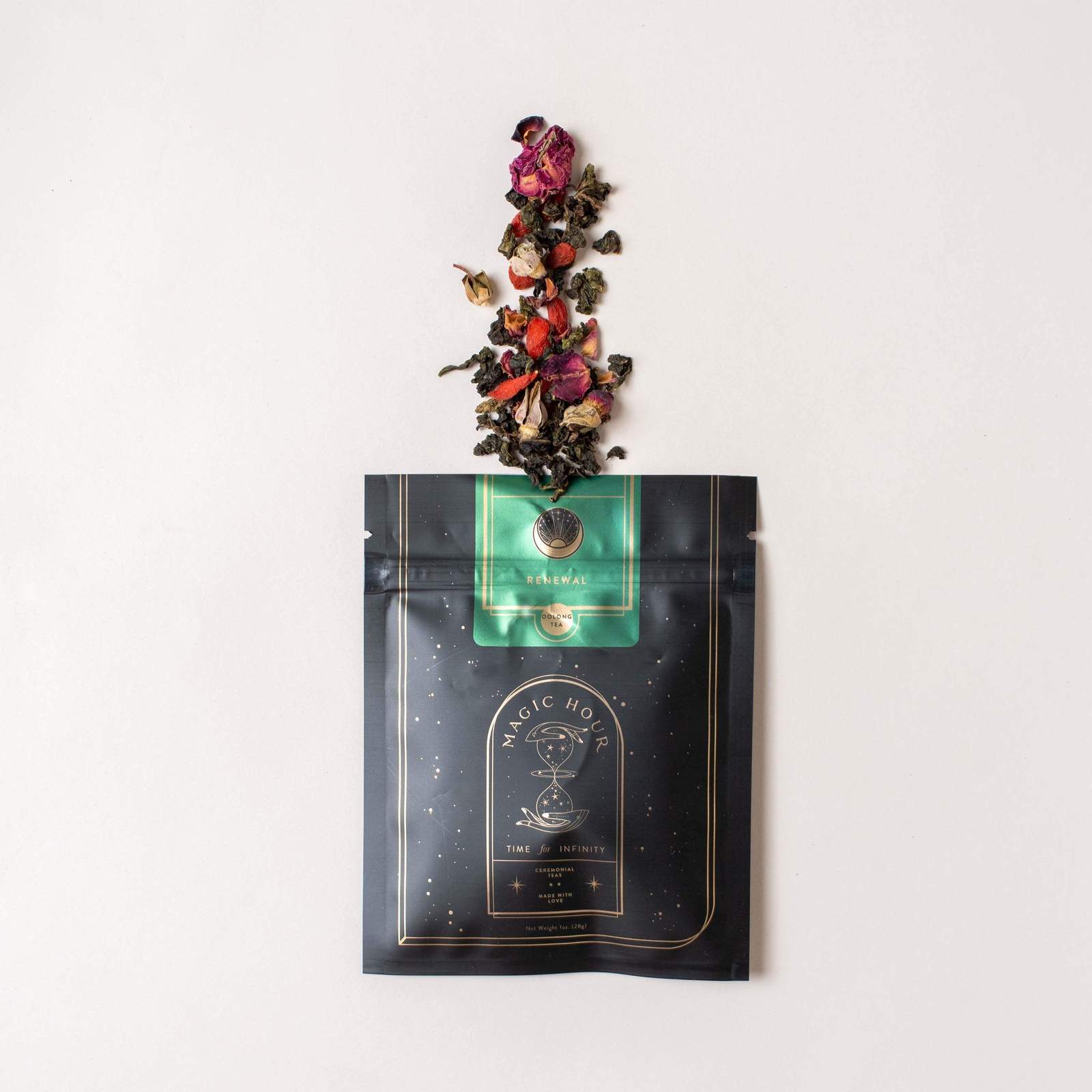 A black and green Magic Hour Renew & Relax: Ritual Gift for Mindfulness & Calm package with intricate gold detailing is laying on a beige surface. The package is open at the top, and a mix of organic loose leaf tea leaves and colorful flower petals is spilling out in a visually appealing arrangement.