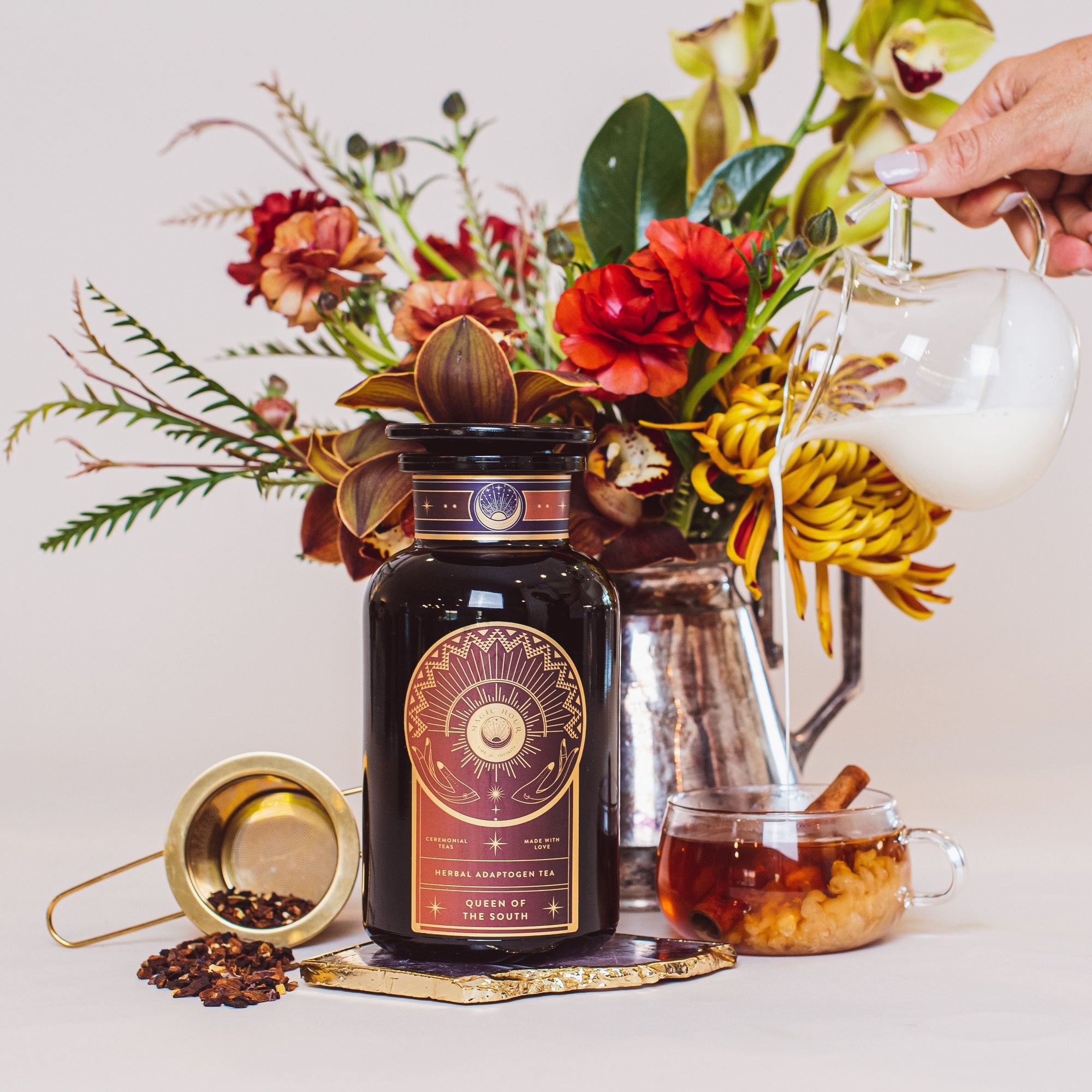 A display featuring a black and gold jar of "Queen of the South: Delicious Cocoa Detox Tea" by Magic Hour, a golden tea strainer with loose tea leaves, a mug of organic tea with cinnamon, a metal pitcher of milk being poured, and a vibrant floral arrangement in the background.