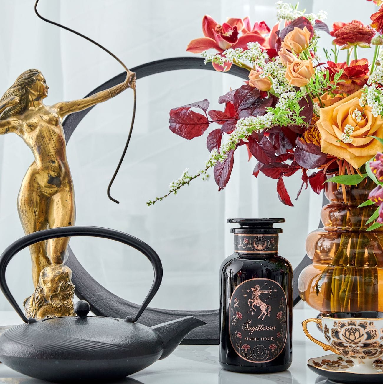 A decorative setup featuring a gold archer statue, a black teapot, a vase with vibrant red, orange, and white flowers, and a black jar labeled "Sagittarius Tea of Good Fortune & Abundance Magic Hour". A teacup and saucer are placed beside the jar, all on a light surface.