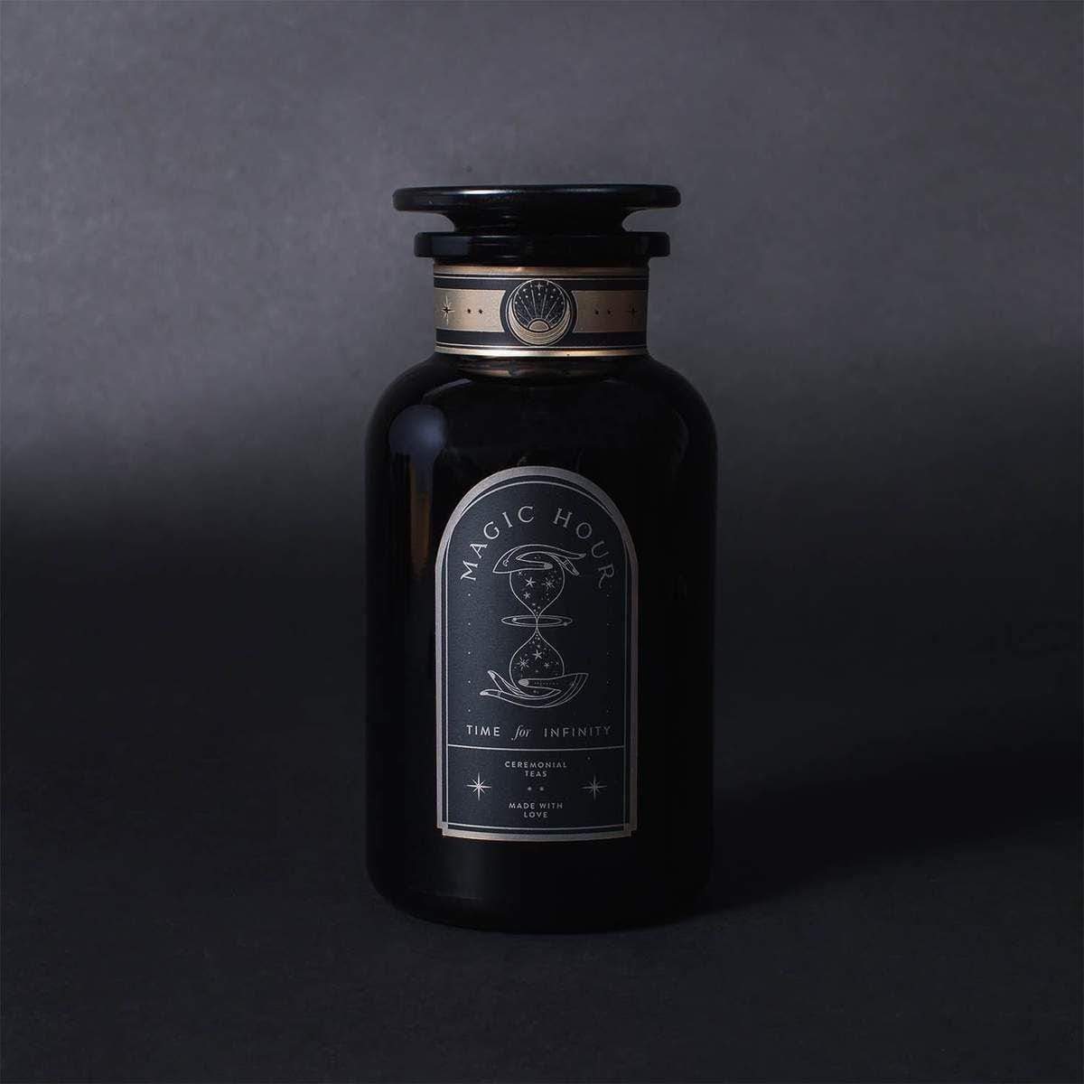 A dark glass bottle with a black label marked "Magic Hour" and illustrations, boasting the text "Time & Infinity." The bottle, reminiscent of Magic Hour's refined tea blend Ti Quan Yin Oolong: Tea of Patient Compassion, has an ornate design and a stopper on top. The background is a gradient of dark gray shades.