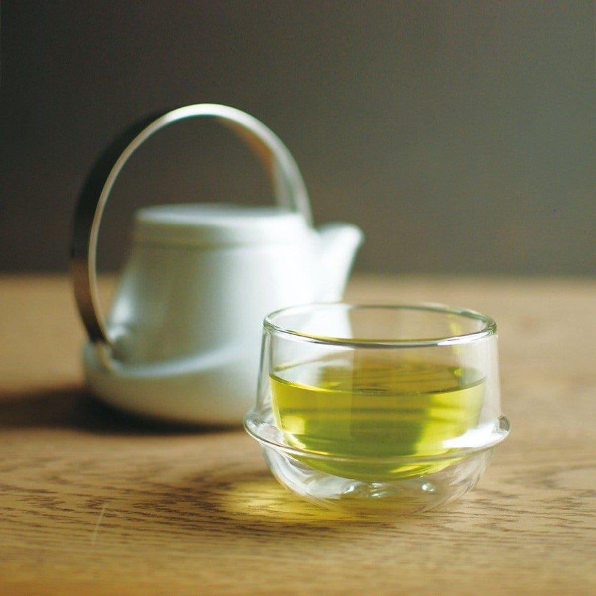 A Kinto Double-Walled Teacup filled with light green loose leaf tea is placed on a wooden surface. In the background, there is a white teapot with a silver handle, slightly out of focus. The setting appears calm and inviting, suggesting a moment of relaxation during the magic hour.