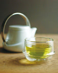 A Kinto Double-Walled Teacup filled with light green loose leaf tea is placed on a wooden surface. In the background, there is a white teapot with a silver handle, slightly out of focus. The setting appears calm and inviting, suggesting a moment of relaxation during the magic hour.
