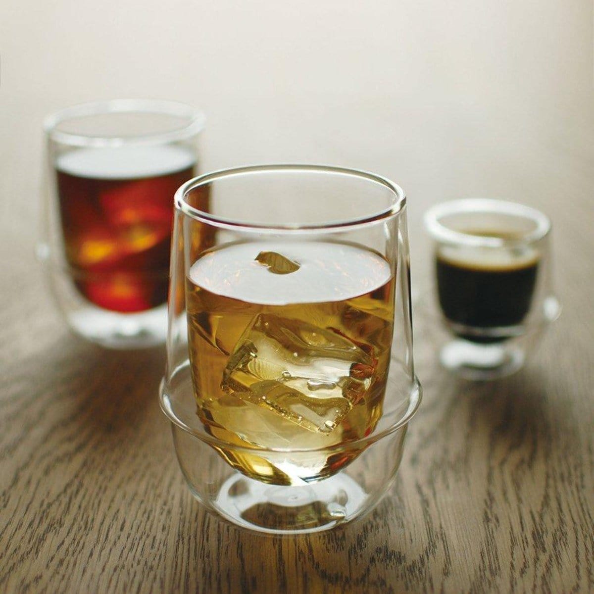 Three double-walled glass mugs are shown on a wooden table. One contains an amber-colored drink with ice cubes, another has a dark brown beverage, and the third, smaller mug holds a dark espresso shot. The amber drink is a refreshing blend of organic tea that perfectly complements the setting in the Kinto Double-Walled Iced Tea Glass: 350mL by Magic Hour.