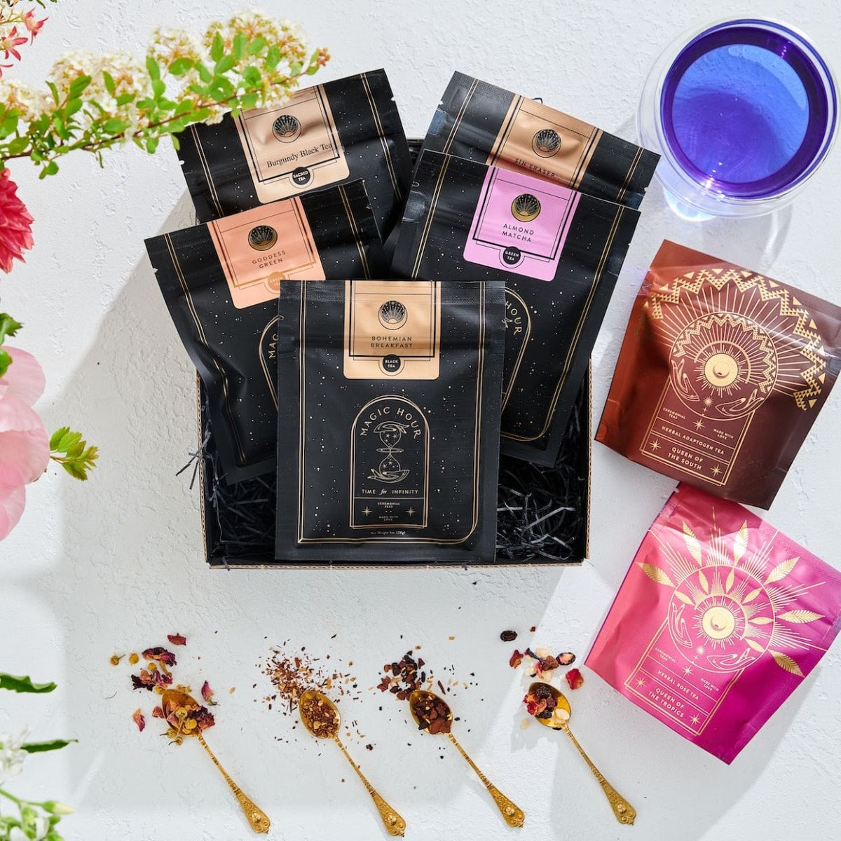 A beautifully arranged flat lay featuring various packets of Bestsellers of Magic Tea Starter Set- 60-75 cup sampler box in black, brown, and pink packaging. Also included are a blue glass, spoons with loose leaf tea and rose petals, and vibrant flowers on a white background, capturing the essence of Magic Hour.