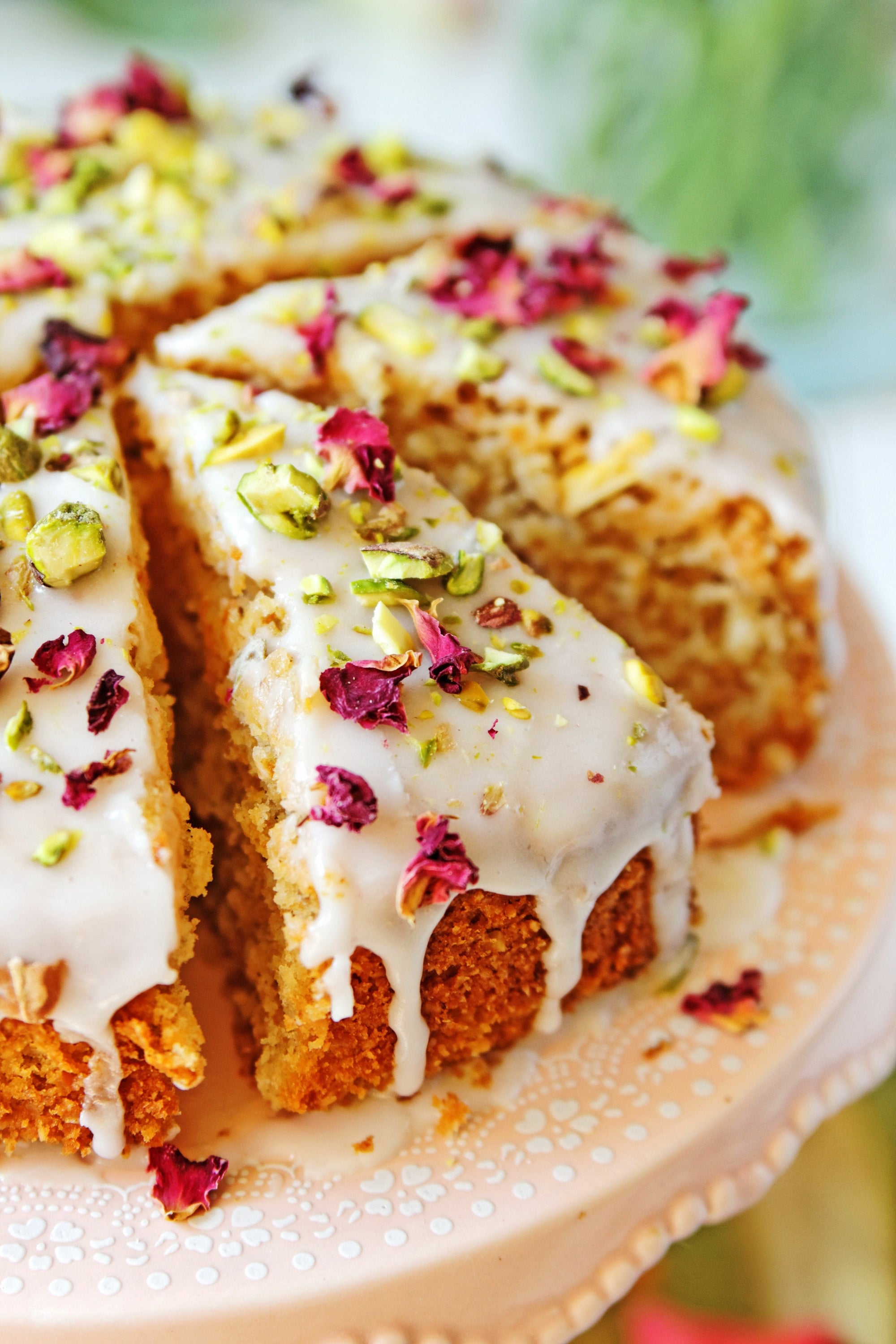 A close-up of a slice of cake topped with glossy white icing, bits of pistachios, and rose petals. The cake sits on a decorative peach-colored plate, with the rest of the cake visible in the background, also topped with icing and garnishes. Enjoy this delightful treat with a cup of Libra- Pistachio-Rose Persian Love Cake Tea with White Pearls & Shatavari by Magic Hour.
