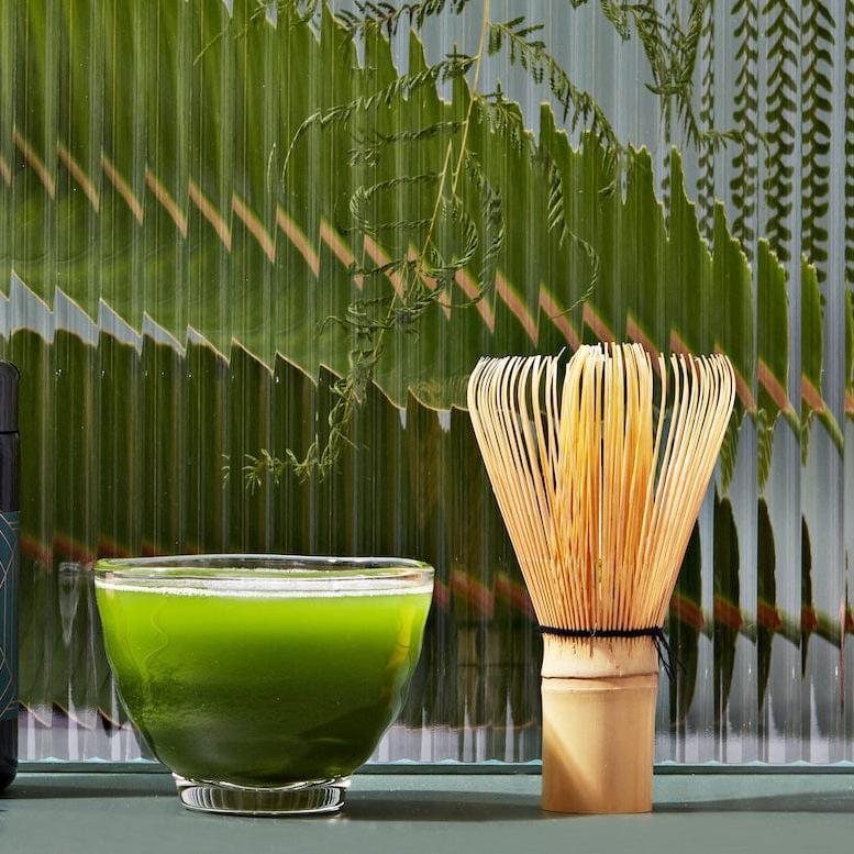 A jar of Magic Hour Kansai-Kyoto Ceremonial Matcha, a bamboo whisk, a small bowl of green matcha tea, and a bamboo scoop are arranged in front of a textured glass panel with green fern leaves visible in the background. The setup is aesthetic and minimalist, highlighting the finest organic tea products.