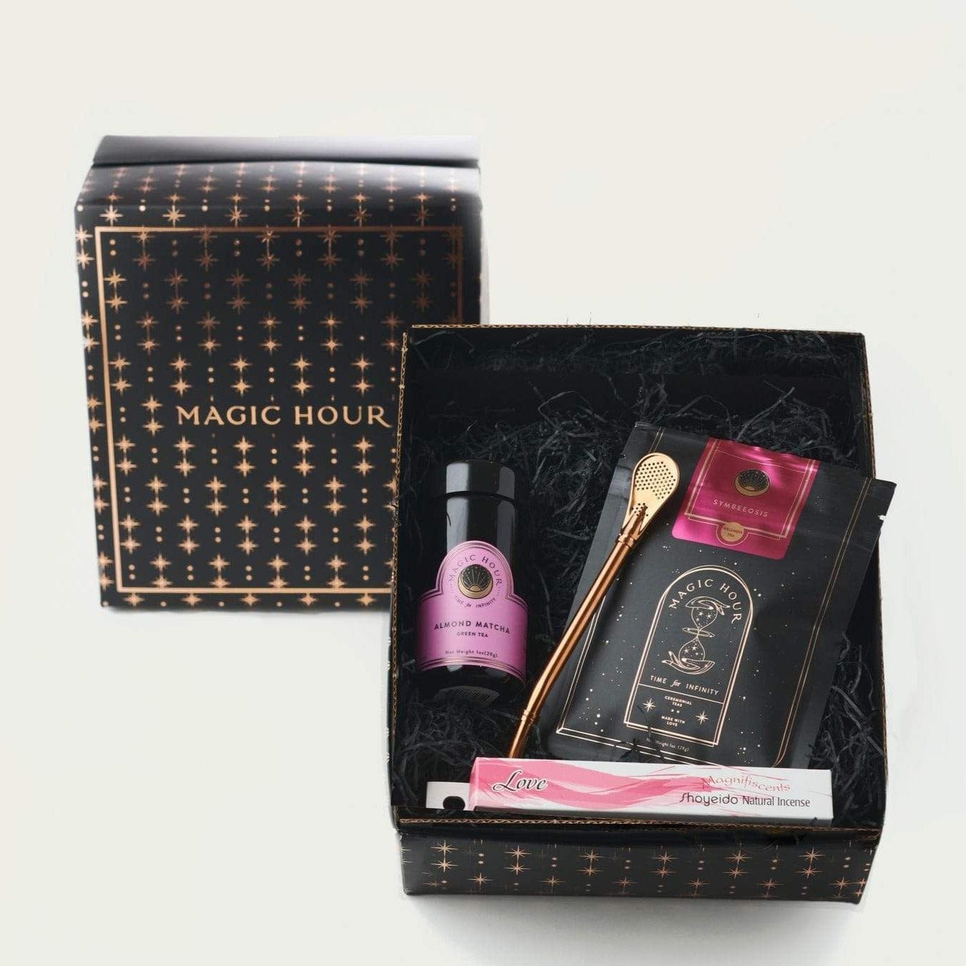 A gift box with "Magic Hour" written on it, containing various items including a small jar with a pink label, a gold spoon, a small glass bottle, a packet of Sip Wellness: Ritual Kit for Immunity & Joy Tea, and a roll of Magic Hour brand incense. The box interior has black shredded paper for padding.