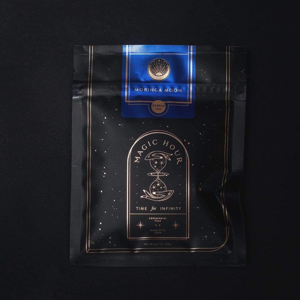 A black package of "Moon Lover: Herbal Immunitea Ritual Gift" organic tea with celestial designs and gold detailing from Magic Hour. The label features a blue section with a tree logo and text, while the main section showcases an hourglass graphic accompanied by "Time for Infinity" and the loose leaf tea type description.