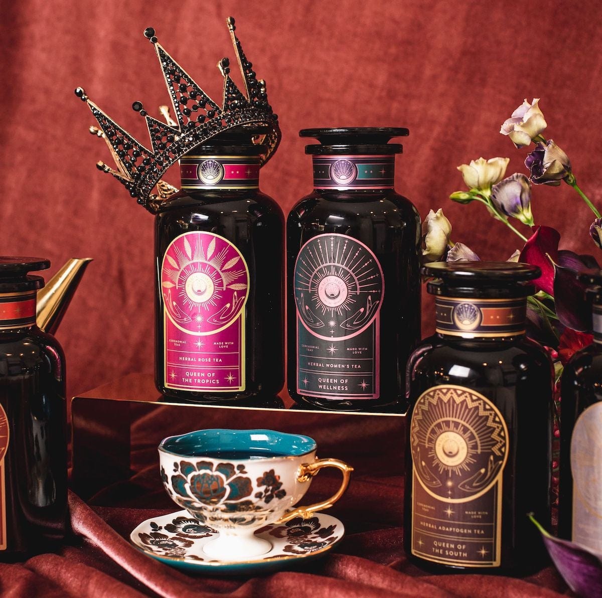 A luxurious display of three ornate tea containers featuring Queen of Wellness: Women&#39;s Hormone Balancing Tea for PMS, Healthy Cycles &amp; Menopause by Magic Hour with regal labels, set against a rich, burgundy backdrop. A decorative crown rests atop one container, and a beautifully detailed teacup and saucer sit nearby. Delicate flowers add a touch of elegance to the scene.