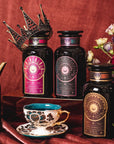 A luxurious display of three ornate tea containers featuring Queen of Wellness: Women's Hormone Balancing Tea for PMS, Healthy Cycles & Menopause by Magic Hour with regal labels, set against a rich, burgundy backdrop. A decorative crown rests atop one container, and a beautifully detailed teacup and saucer sit nearby. Delicate flowers add a touch of elegance to the scene.
