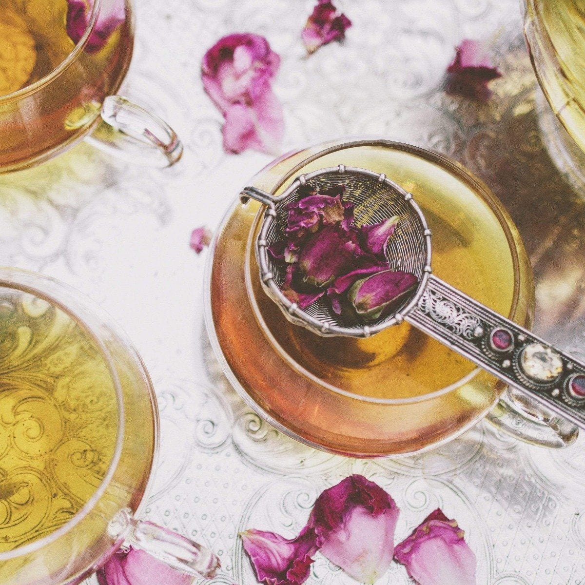 A close-up of three tea cups filled with light brown Wild Indian Roses by Magic Hour, placed on an ornate silver tray. Rose petals are scattered around and one silver strainer filled with rose petals rests on top of one cup. The scene has a delicate, elegant feel, embodying the essence of fine Organic Tea.